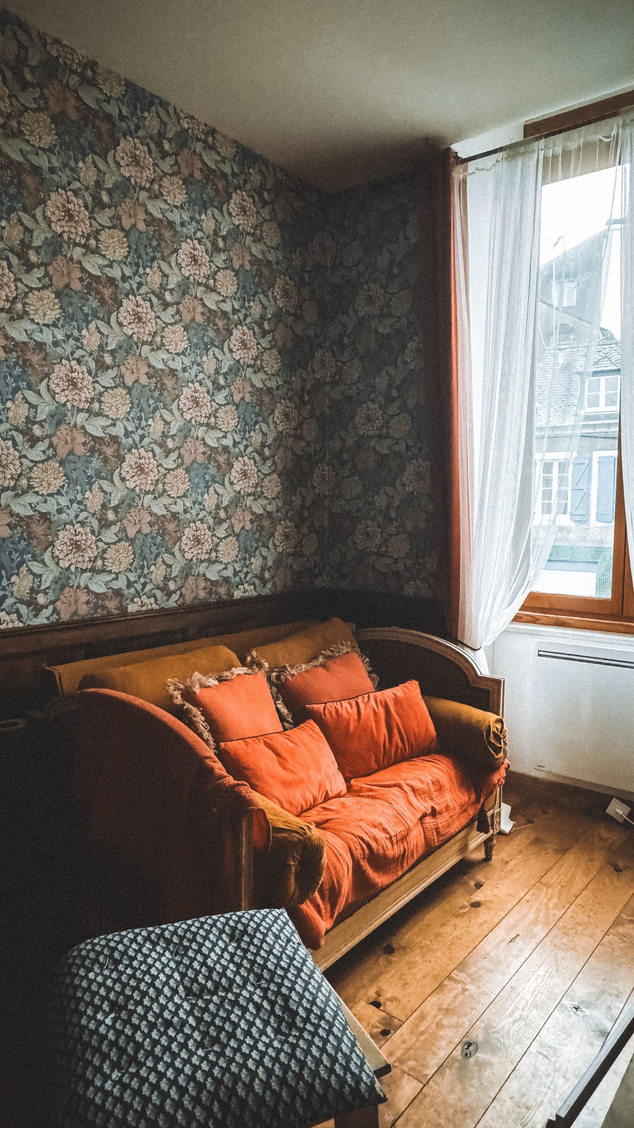 Repeating floral wallpaper in a sitting room with an orange sofa beside a sunlit window