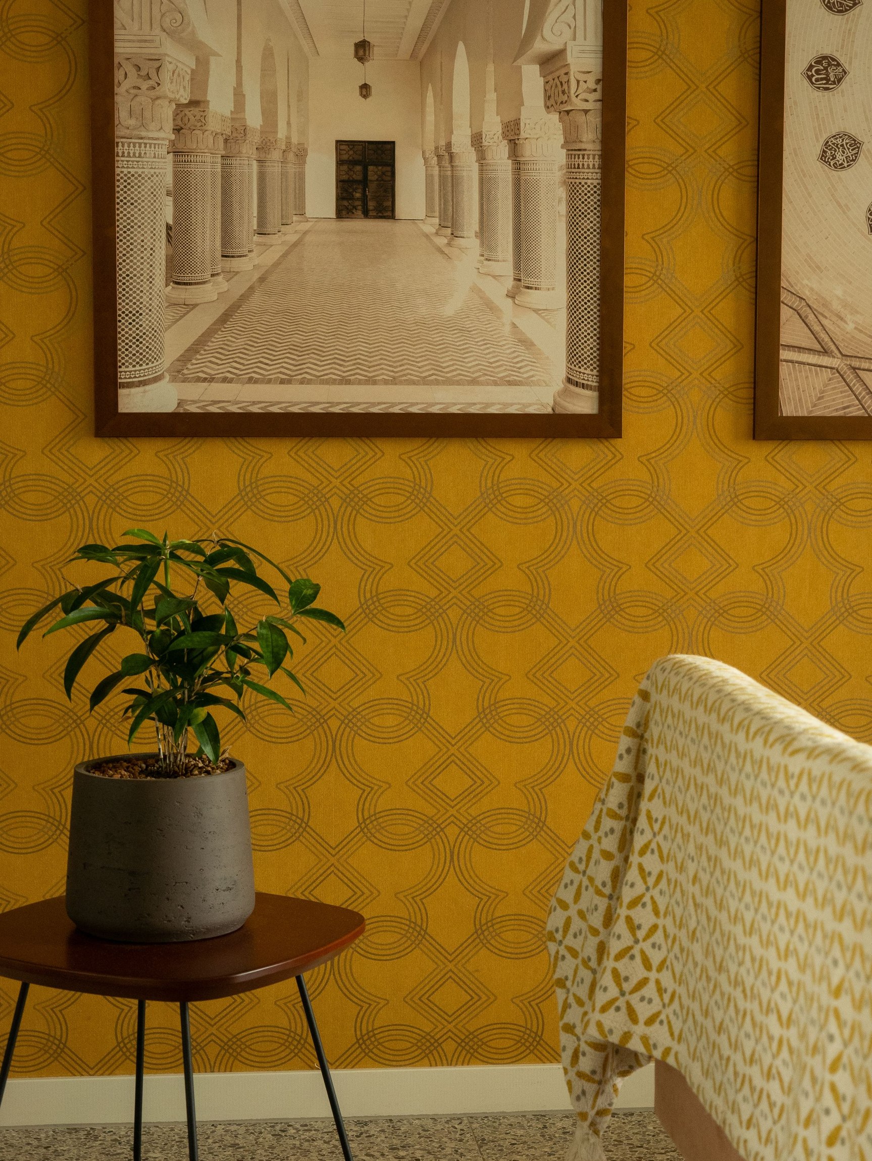 Gold repeating geometric wallpaper behind framed artwork, a plant and a chair